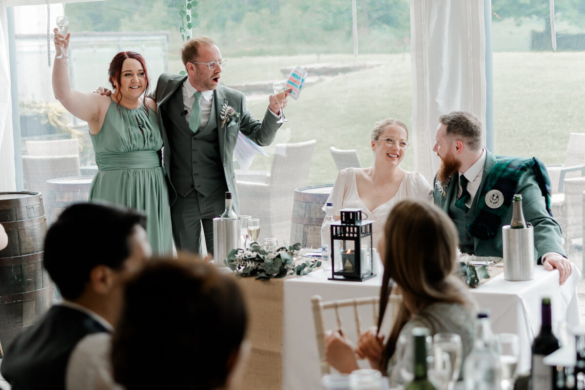 Wedding guests raising champagne glasses during a lively speech as the bride and groom laugh at their reception at Ardoch.