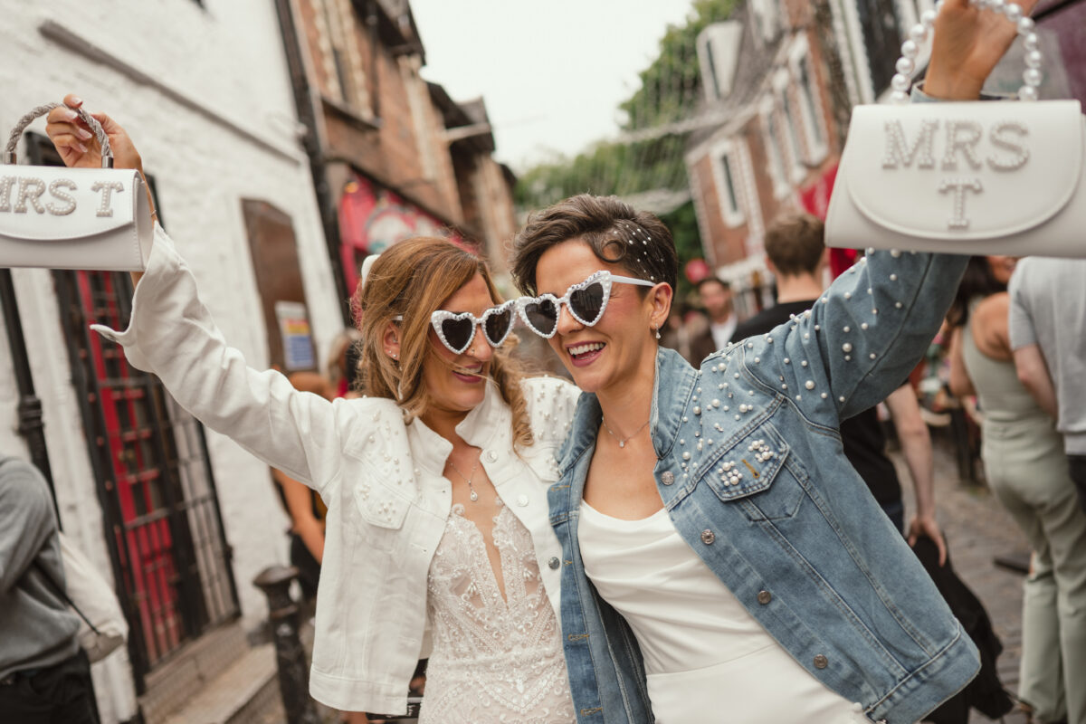 Two brides laughing together in heart-shaped sunglasses and holding personalised bridal handbags during their Glasgow West End wedding celebrations in Ashton Lane