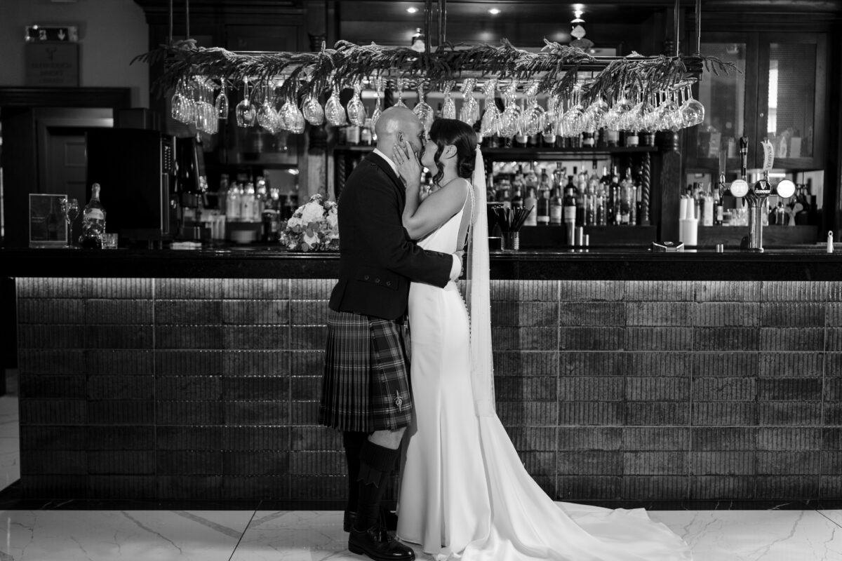 Black and white wedding photograph of a bride and groom kissing in front of the bar.