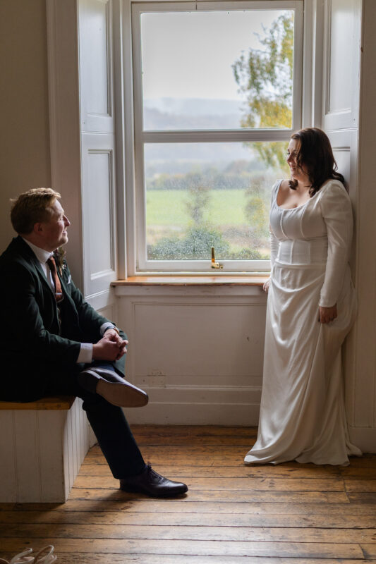 Bride in a long-sleeved satin wedding dress smiling at her groom in a dark green suit beside a large rain-speckled window at Comrie Croft in Perthshire.