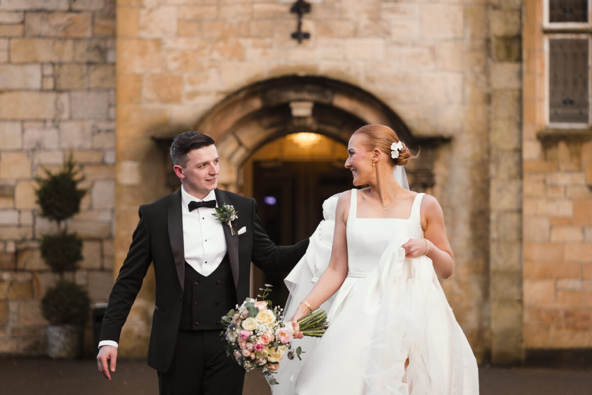 Bride holding a pastel rose bouquet smiling at her groom in a black tuxedo outside the stone entrance of Dalmeny Park.