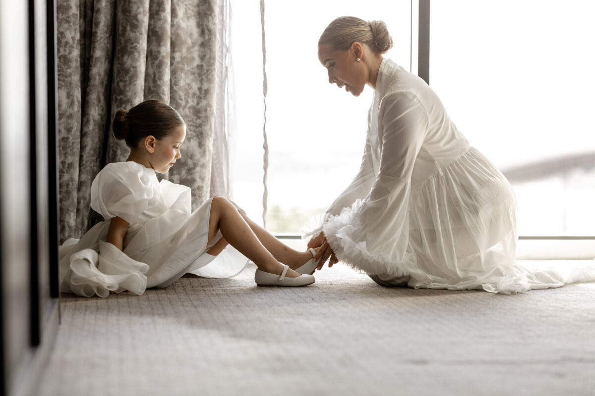 Bride helping a young flower girl with her white shoes beside a window during wedding morning preparations.