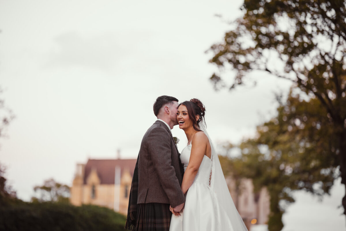 Bride laughing as her groom leans in close to whisper in her ear during a romantic outdoor wedding portrait at Glenbervie House, with the historic venue in the background.