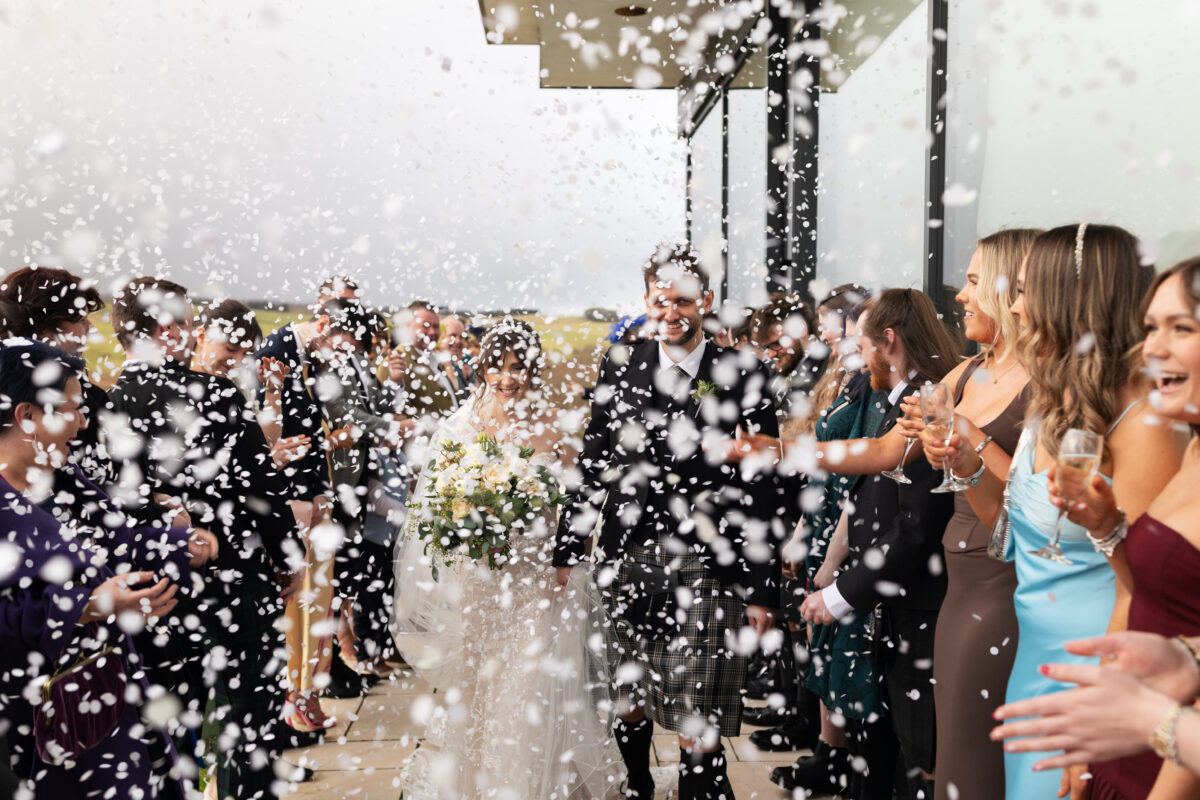 Bride and groom walking hand in hand through a shower of white confetti as wedding guests celebrate outside Lochside House Hotel, with the groom wearing a traditional Scottish kilt.