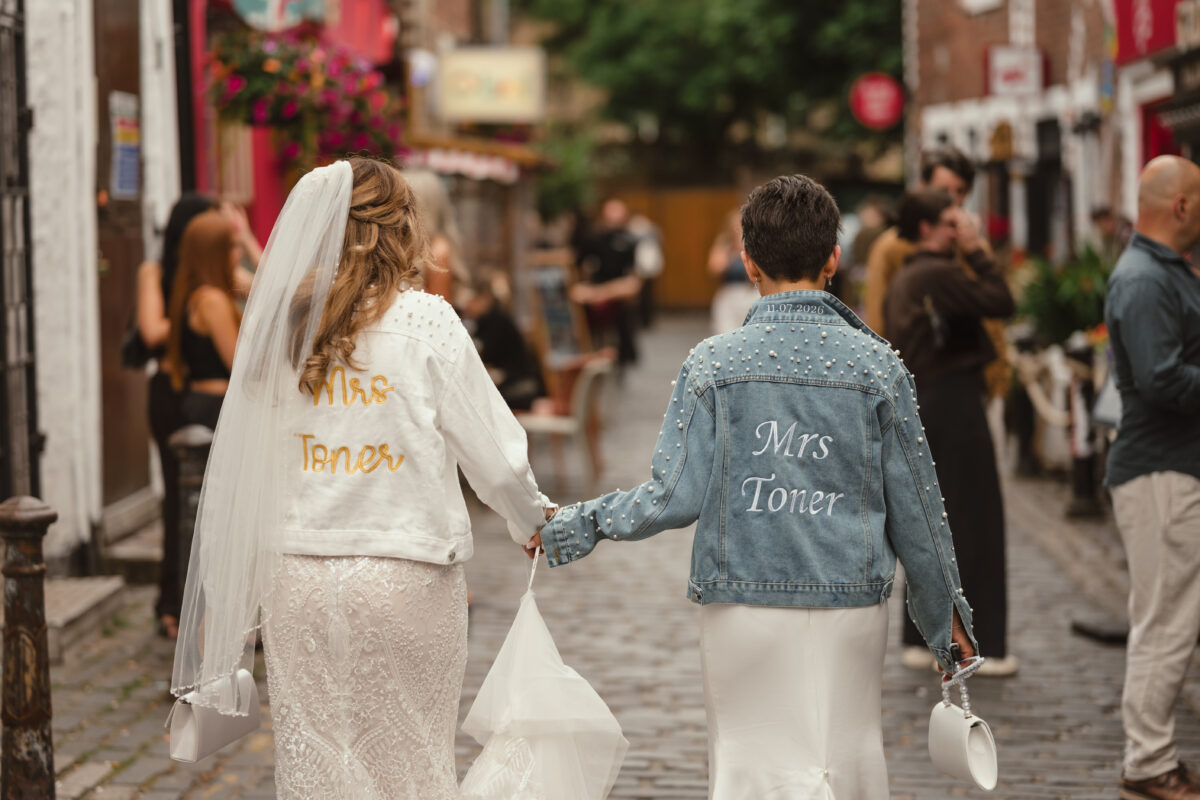 Two brides walking hand in hand along a cobbled street, Ashton Lane, in Glasgow's West End, wearing personalised Mrs Toner wedding jackets.