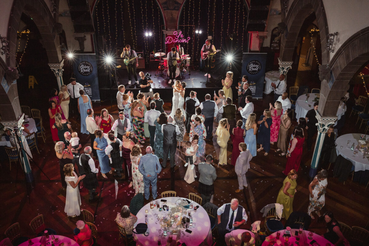 Wedding guests celebrating on the dance floor beneath the grand stone arches of Òran Mór in Glasgow, with a live wedding band performing.